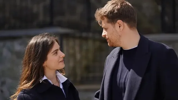 Two adults conversing outdoors, facing each other; woman has dark hair and white collared shirt, man wears a dark jacket.