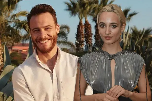 Man with a beard and a woman with blonde hair posing together outdoors, smiling, with palm trees in the background. He wears a white shirt; she wears a metallic gray dress.