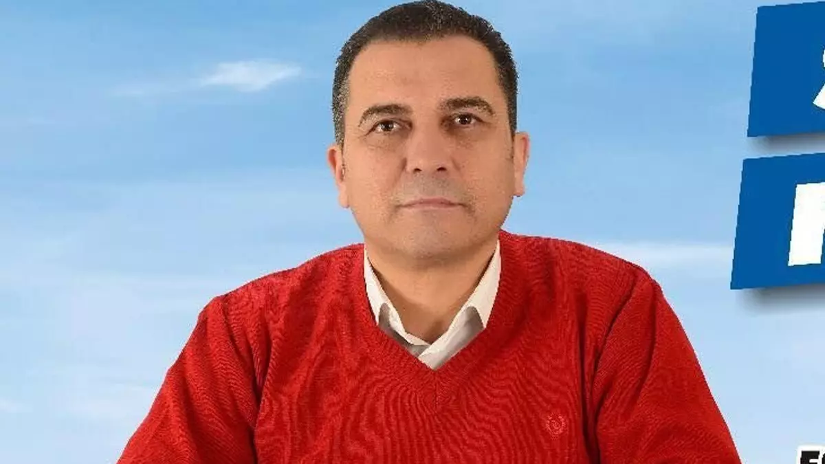 Man in a red sweater sits facing the camera against a light blue background with white collar showing, looking at the viewer.