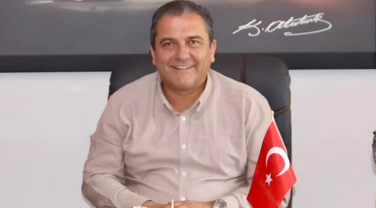 Smiling man in a light pink shirt sits at a desk in an office, Turkish flag on his right side.