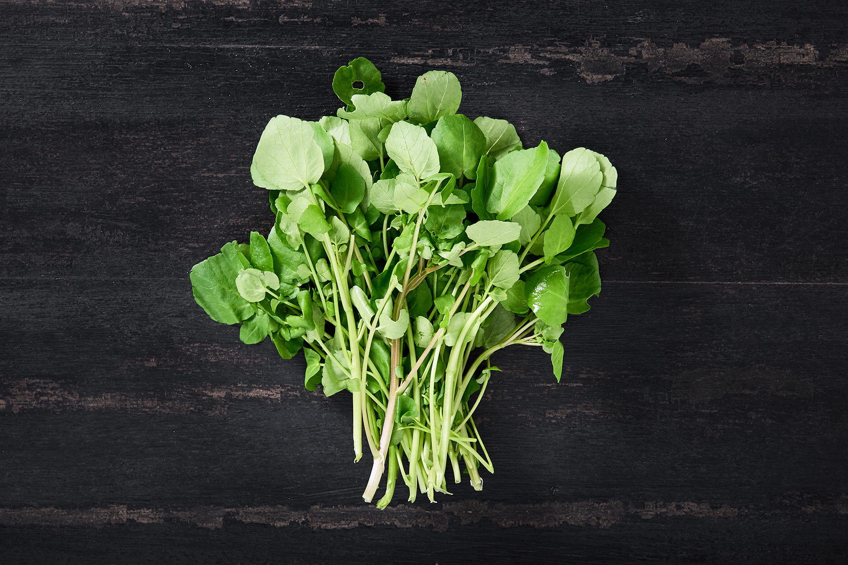 Bundle of fresh watercress with slender stems and rounded green leaves on a dark wooden table surface