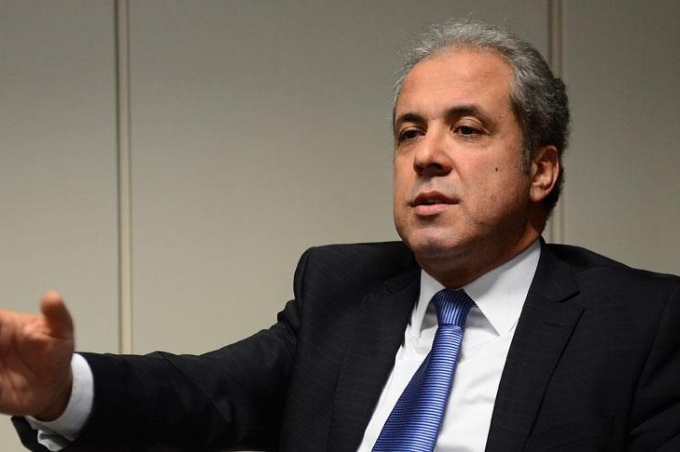 Businessman in a dark suit and blue tie speaks and gestures during a meeting or interview.