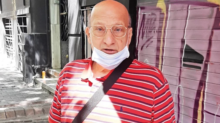 Older man with glasses, striped red shirt, and a face mask under his chin stands on a sidewalk with graffiti behind him.