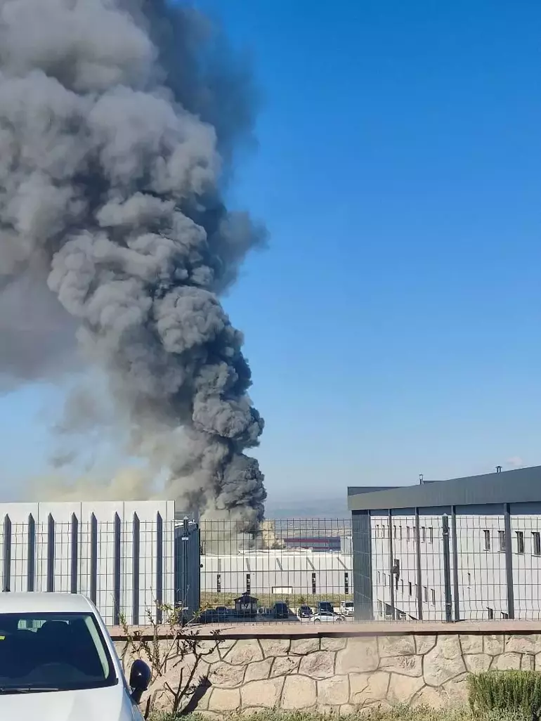 Industrial facility emitting a tall column of dark smoke into a clear blue sky, with a fence and parked cars in the foreground.