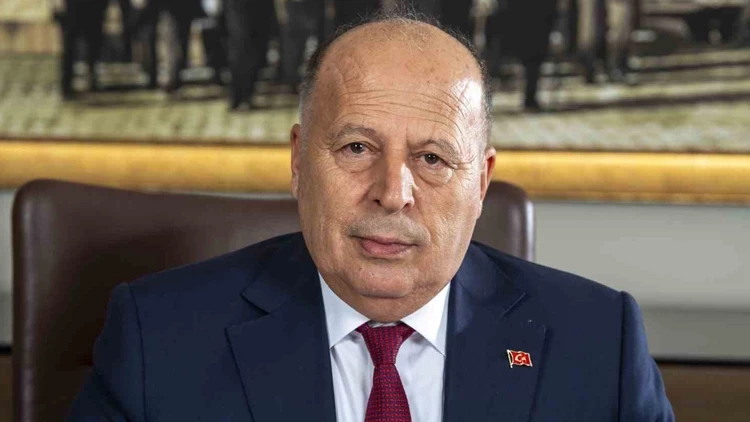 Older man in a navy suit and red tie sits at a desk, wearing a Turkish flag lapel pin.