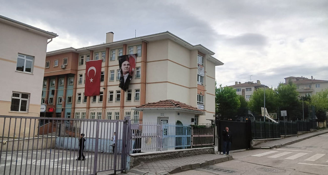 School building with a Turkish flag and an Atatürk banner on the facade, a fenced yard in front and pedestrians nearby.