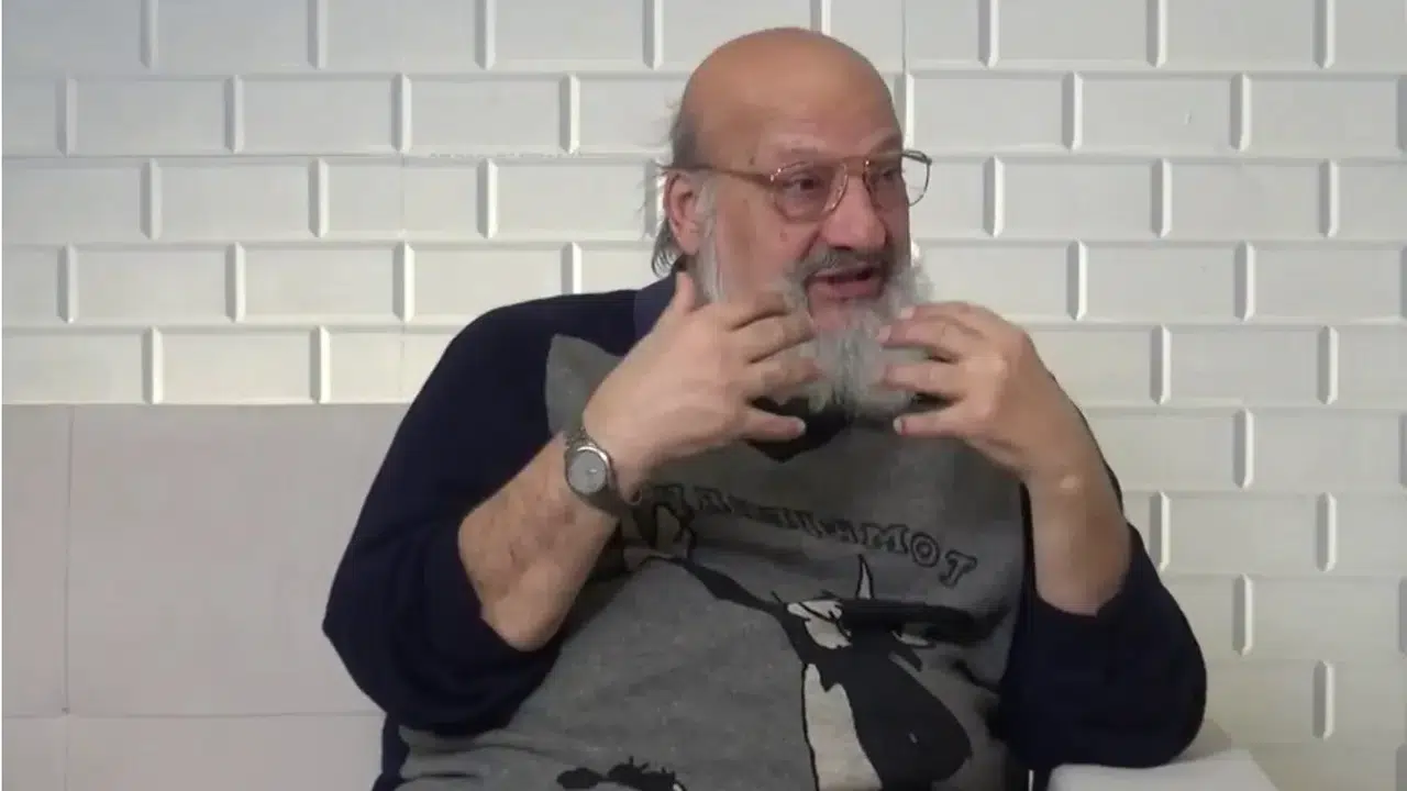 Older man with glasses and a gray beard gesturing with his hands while speaking, seated against a white textured wall.