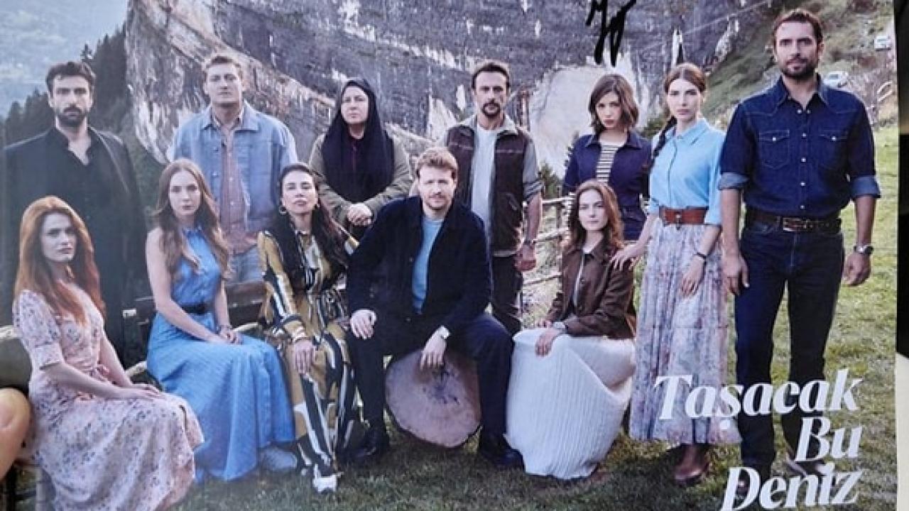Group of ten adults posing outdoors on a grassy hillside with rocky cliffs in the background; everyone dressed stylishly for a photo, some seated and others standing. A title overlay reads 'Tasacak Bu Deniz' in the corner.