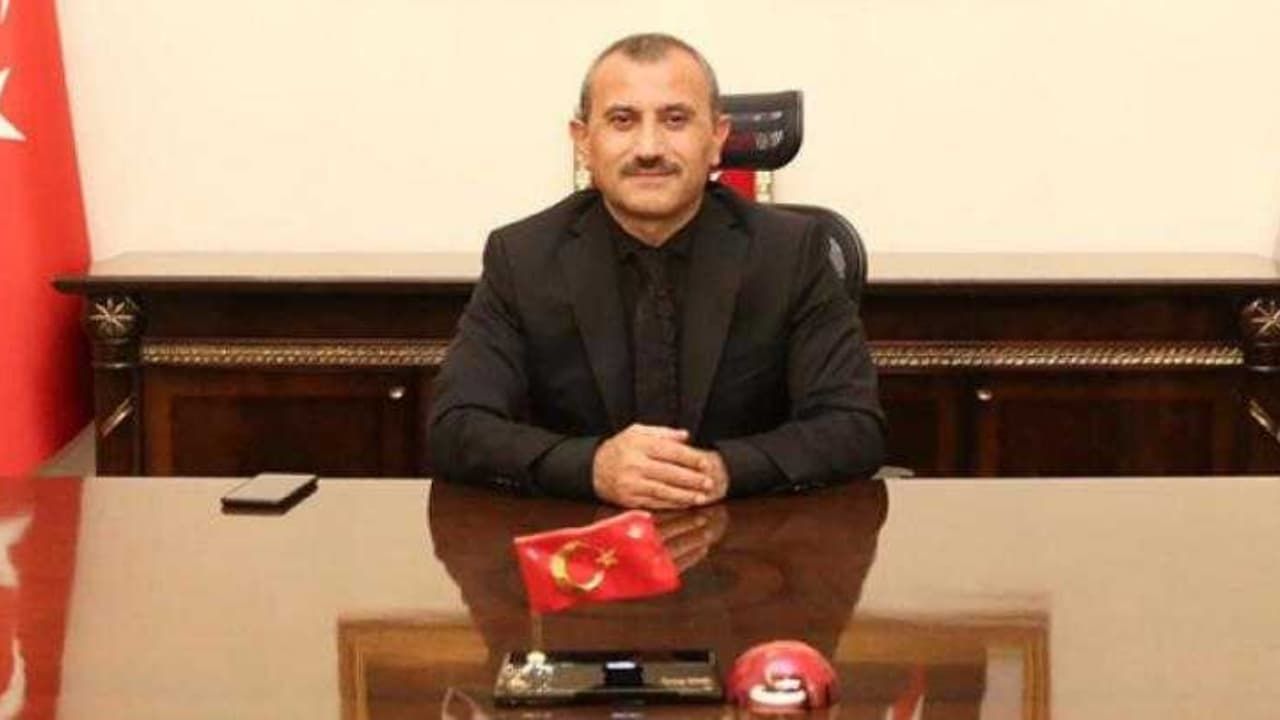 Man in a black suit seated at a polished desk in an office, with a small Turkish flag on the desk in front of him.